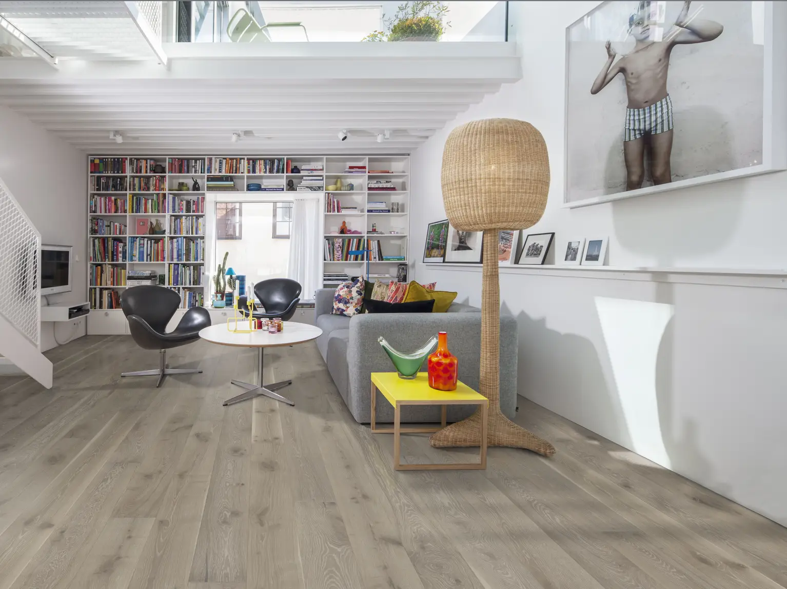 Oak nouveau. Modern living room with wooden floors, a gray sectional sofa, two black chairs, a large bookshelf, and a tall woven floor lamp.