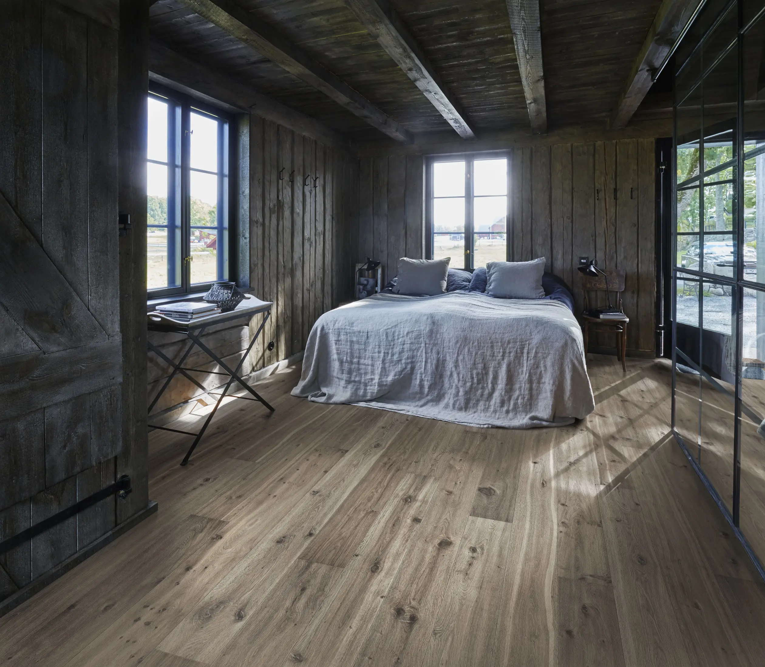 Cozy rustic bedroom with wooden walls and floor, featuring a double bed with gray bedding, large windows, and soft natural light.