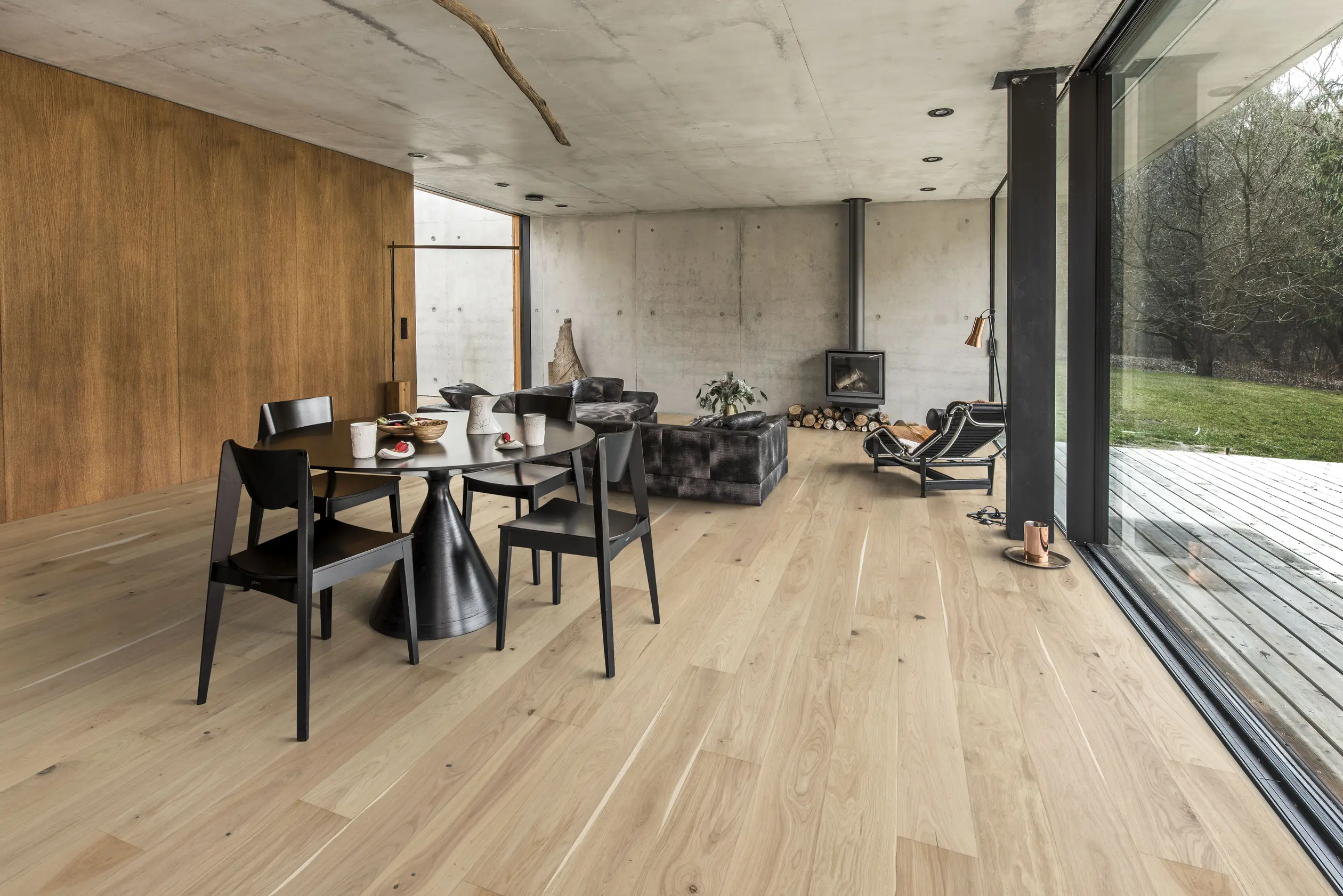 Modern living and dining area with wooden floors, concrete walls, black furniture, large windows, and a view of a grassy outdoor area.