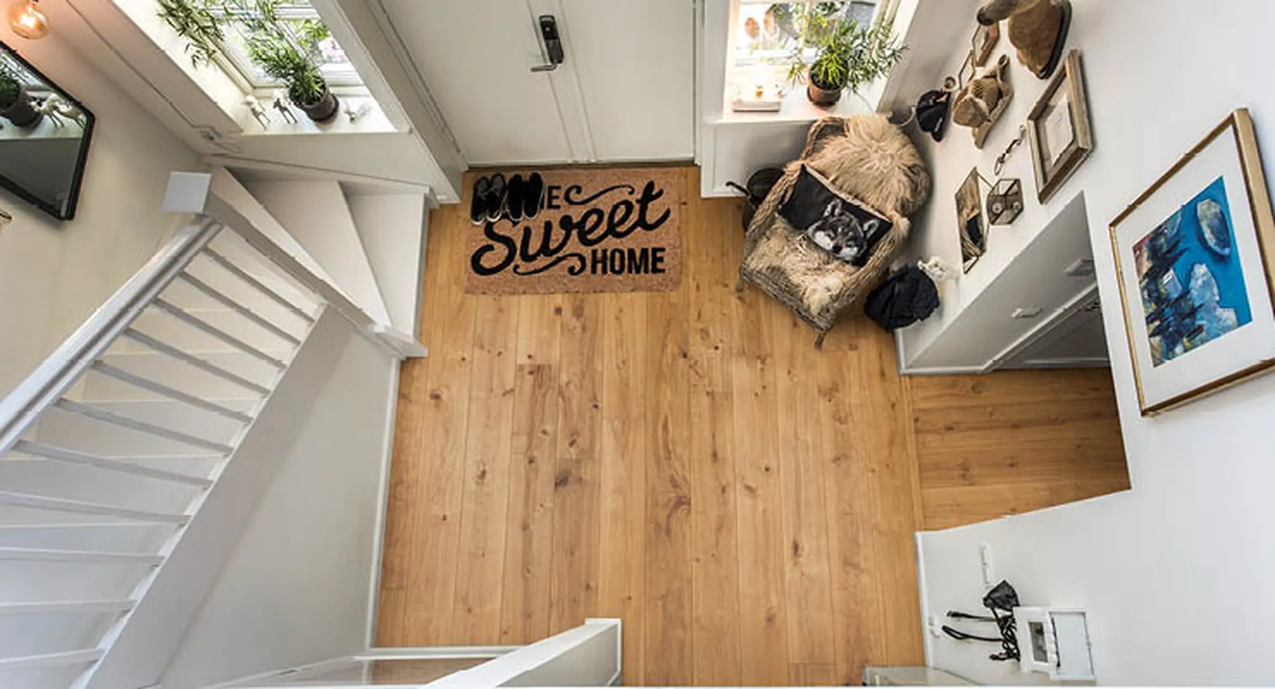 A cozy, well-lit entryway with a "Home Sweet Home" doormat, wooden floor, stairs, and wall decor, including plants and framed pictures.