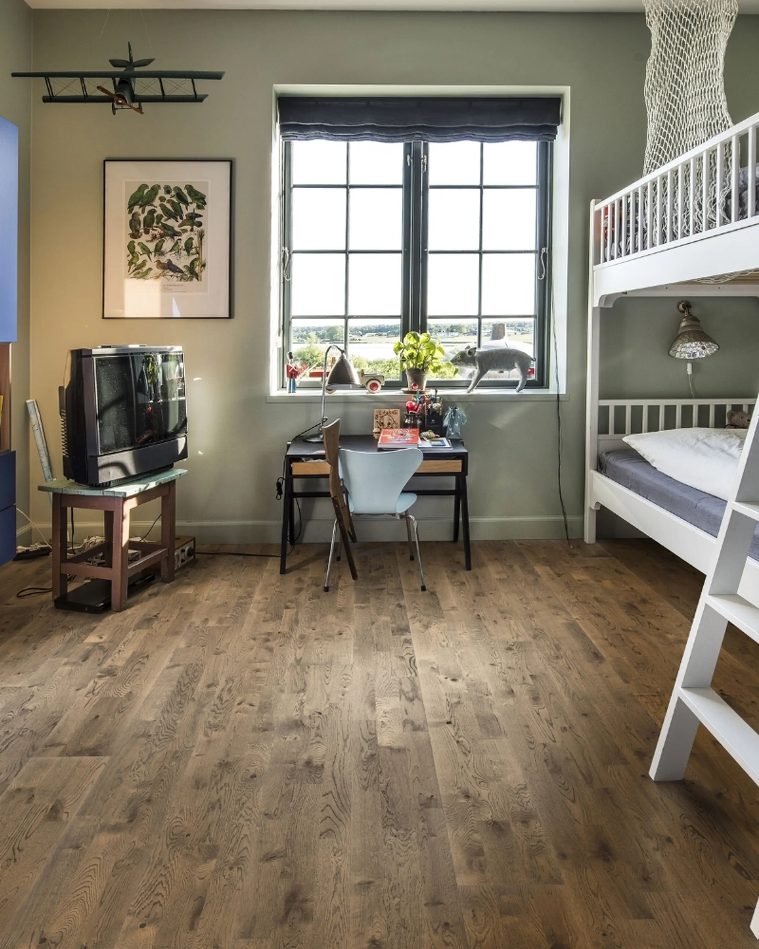 A bright children's room with mid-tone wooden flooring, bunk beds, a desk by the window, and playful decorations like a hanging airplane model.