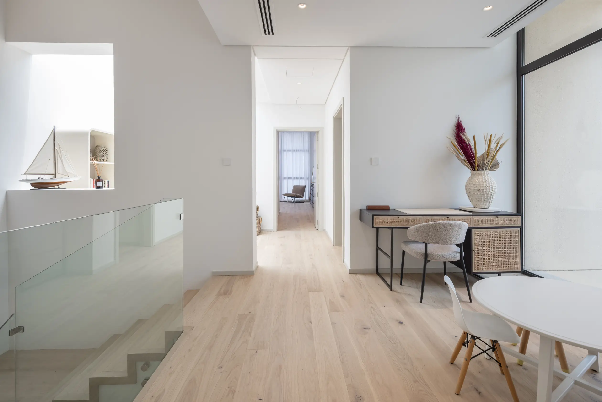 Modern interior with light wood flooring, white walls, a desk with a vase, and a small round table with chairs, leading to a bright hallway.