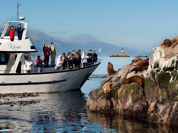Boat tour passengers observe sea lions on rocks in Ushuaia National Park with mountain backdrop.