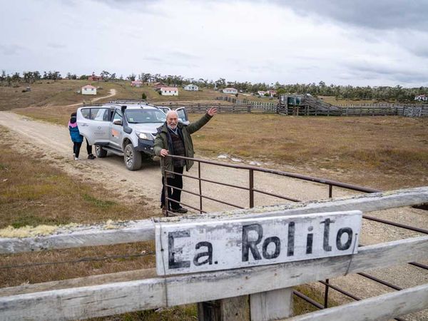 Man gesturing at La Rolita gate on rural Patagonian estancia with white vehicle and ranch buildings in background.