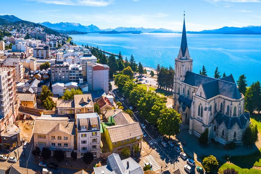 Aerial view of Bariloche's lakefront cathedral and colorful residential buildings overlooking turquoise Lago Nahuel Huapi with snow-capped Andes mountains.