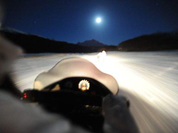 Moonlit winter landscape with snow-covered terrain, mountains silhouetted against night sky, and vehicle headlights illuminating Patagonian wilderness.