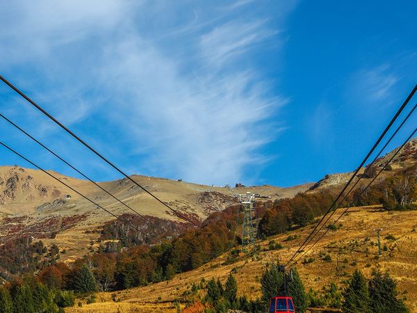 Red cable car ascending mountain slope near Bariloche with autumn forest and snow-capped peaks under blue sky.