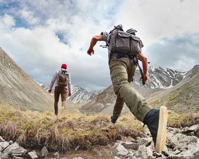 Hikers trekking through a rocky alpine valley with snow-capped mountains and dramatic terrain under cloudy skies.