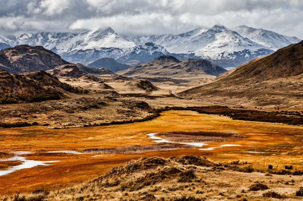 Snow-capped peaks rise above a golden wetland valley with mineral-orange streams in Patagonia's Carretera Austral region.