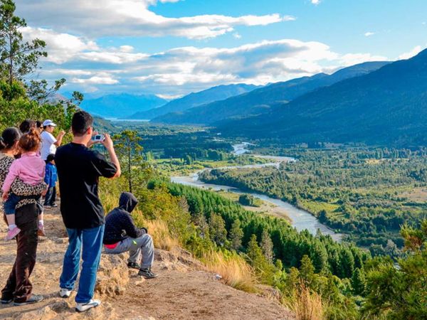 Tourists photograph a sweeping valley view with a winding river, forested mountains, and blue sky near El Bolsón.