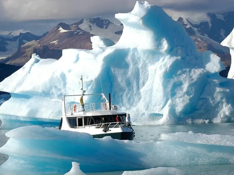 Boat sailing past huge blue icebergs on a lake, with snow-covered mountains in the distance and a cloudy sky. Los Glaciares National Park