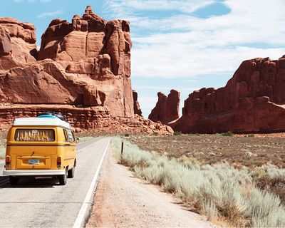Yellow campervan drives along a desert highway surrounded by striking red rock formations and sparse vegetation.
