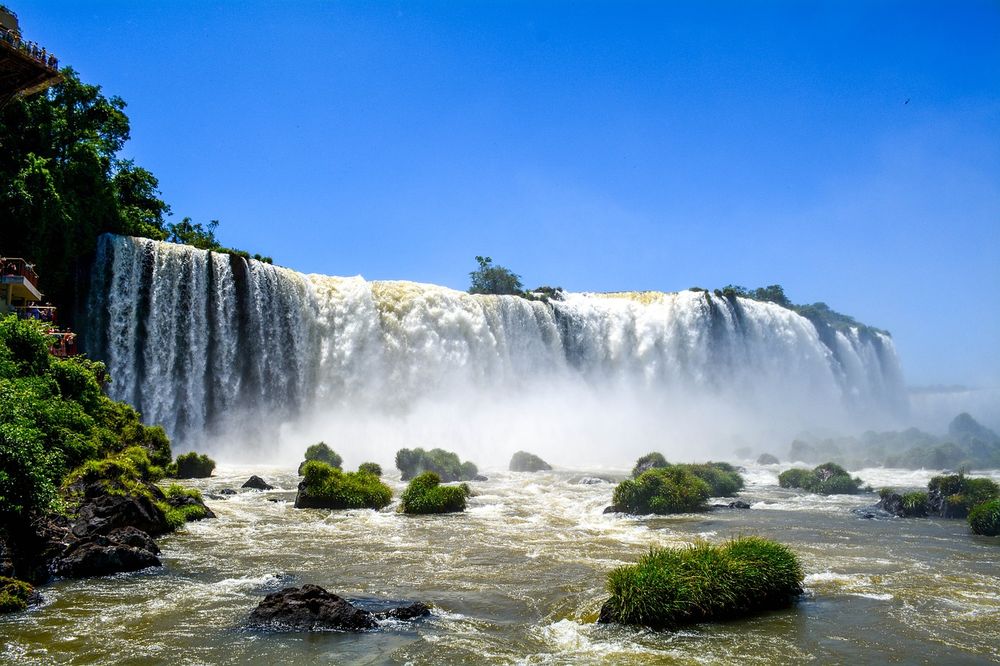 Massive cascading waterfalls of Iguazú with mist rising above the turbulent river and lush green vegetation framing the dramatic landscape.