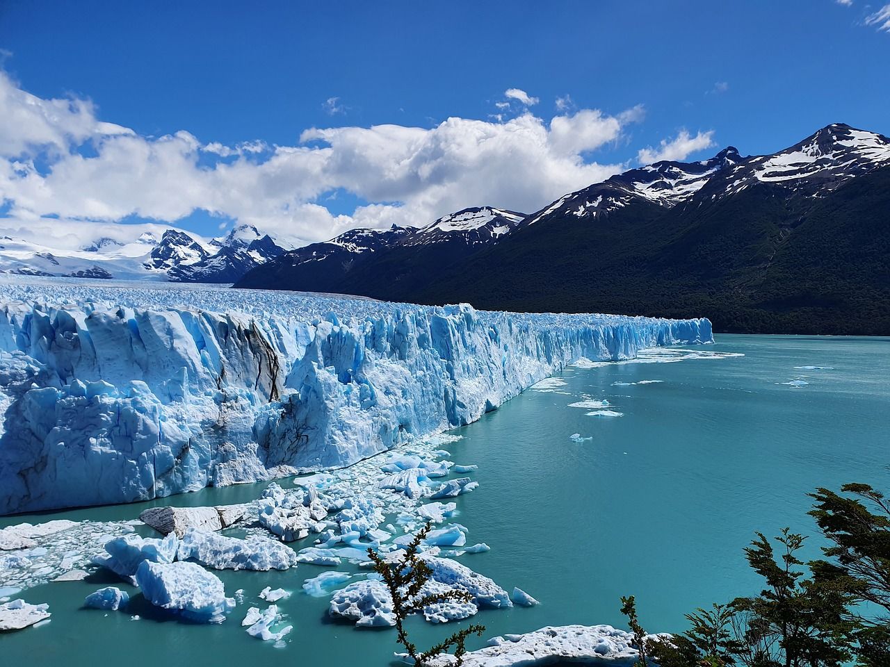 Glaciar Perito Moreno's massive blue ice wall meets turquoise waters with snow-capped mountains under clear skies in El Calafate.