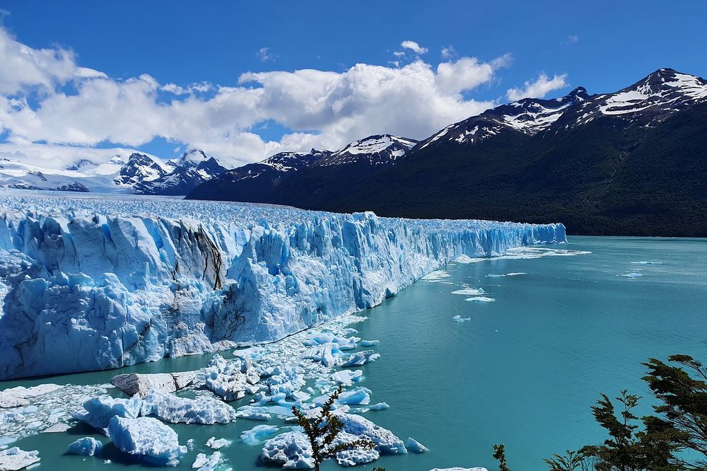 Glaciar Perito Moreno's massive blue ice wall meets turquoise waters with snow-capped mountains under clear skies in El Calafate.