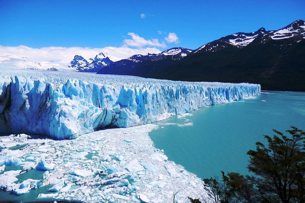 Glaciar Perito Moreno's towering blue ice formations meet turquoise meltwater beneath snow-capped Patagonian peaks under clear skies.