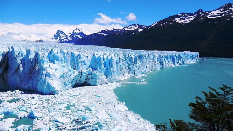 Glaciar Perito Moreno's towering blue ice formations meet turquoise meltwater beneath snow-capped Patagonian peaks under clear skies.
