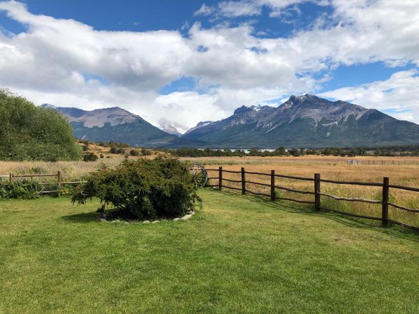Green lawn, wooden fence, golden field, lake, and majestic snow-capped mountains under a partly cloudy sky. Nibepo Aike Estancia