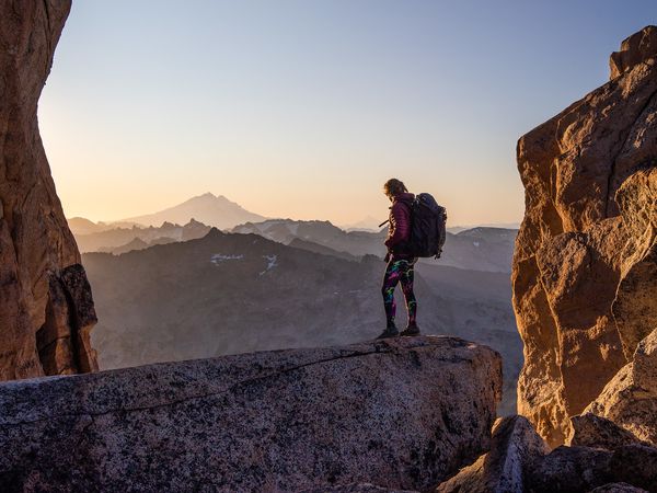 Hiker with backpack stands on rocky outcrop at sunrise near Mount Tronador, Bariloche, with misty mountains in background.