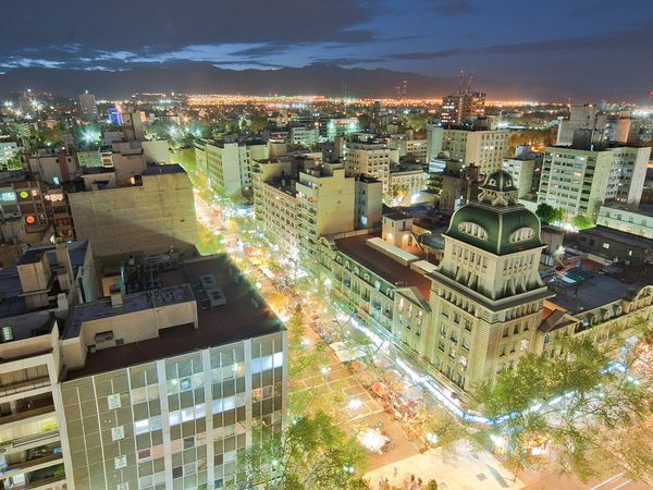 Mendoza city skyline illuminated at dusk with tree-lined streets, historic architecture, and Andes mountains in the distance.