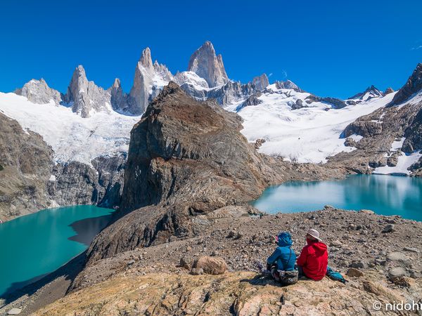 Two hikers rest above turquoise glacial lakes with Mount Fitz Roy's granite peaks and snow-covered slopes in El Chaltén, Patagonia.