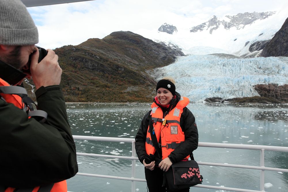 Tourist photographing glacier landscape from cruise ship deck in Patagonia, wearing orange life vest.