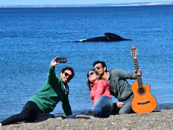 Three travelers pose with a guitar on Doradillo Beach in Puerto Madryn while a whale surfaces in the background.
