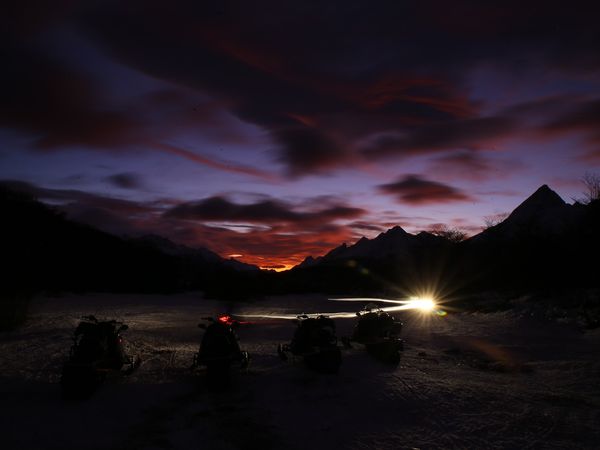 Campfire glowing beside a Patagonian river at twilight, with snow-capped mountains silhouetted against a dramatic sunset sky.