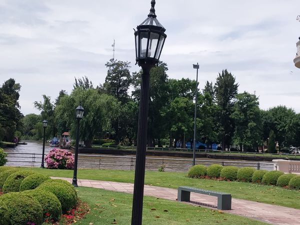 Elegant riverside park in Tigre, Buenos Aires, with ornate black street lamp, manicured gardens, and calm waterway reflections.
