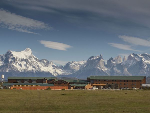 Red ranch buildings with green roofs stand on vast Patagonian grassland beneath dramatic snow-capped Andes peaks near Río Serrano.