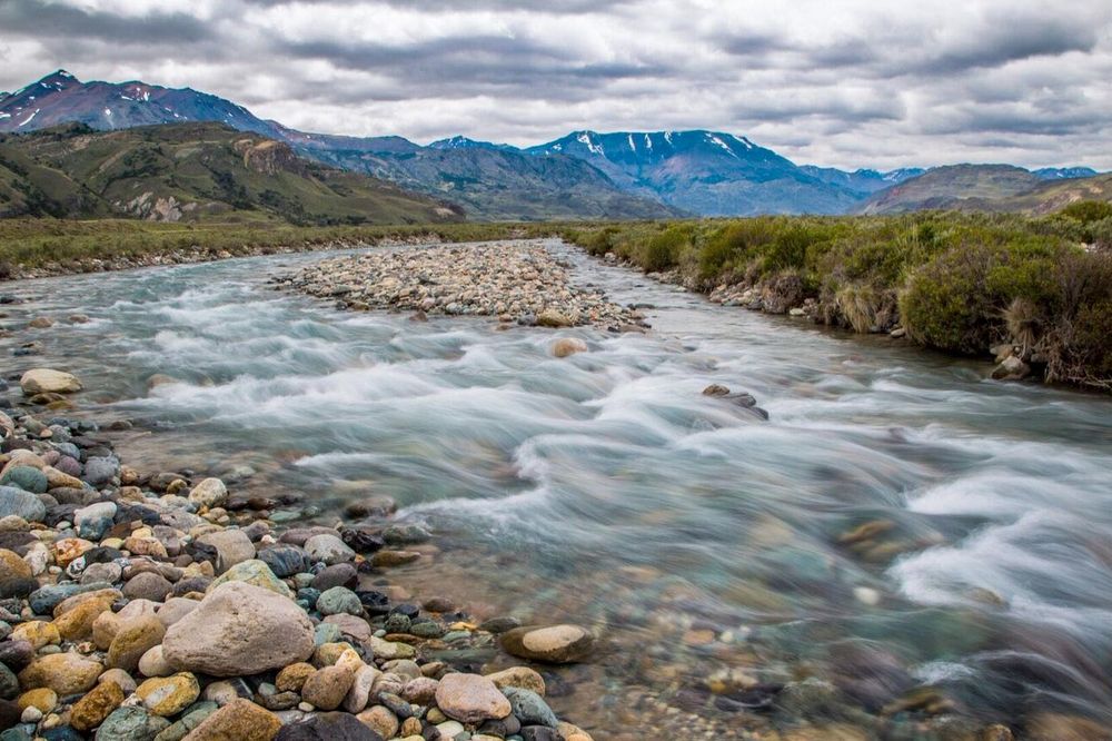 Turquoise river flows through rocky terrain with snow-capped mountains and Patagonian steppe vegetation under cloudy skies.