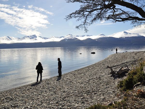 Visitors explore the pristine pebbled shores of Lago Escondido with snow-capped Patagonian mountains reflected in calm waters.
