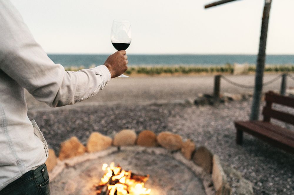 Person holding red wine by a beach bonfire at Bahía Bustamante, Patagonia, with ocean and rocks in background.