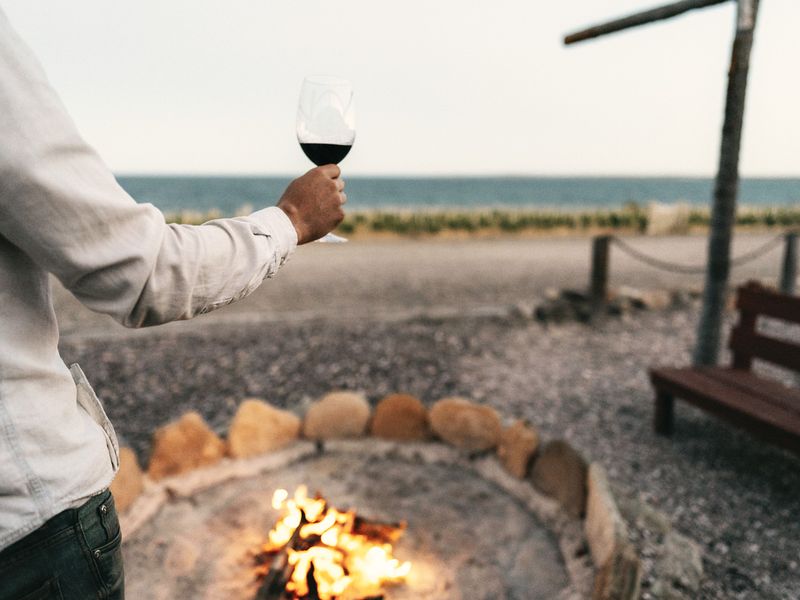 Person holding red wine by a beach bonfire at Bahía Bustamante, Patagonia, with ocean and rocks in background.