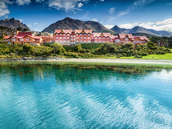 Colorful red hotel buildings nestled among green vegetation along turquoise river waters with dramatic mountains in background.