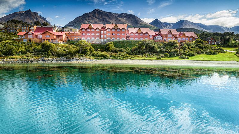 Colorful red hotel buildings nestled among green vegetation along turquoise river waters with dramatic mountains in background.