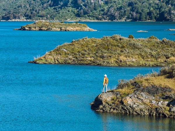 Hiker in yellow jacket stands on rocky cliff overlooking turquoise waters and forested islands in Tierra del Fuego National Park.