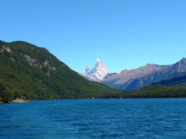 Vast blue lake with forested shores, leading to jagged, snow-capped mountains like Fitz Roy under a bright blue sky. Lago del Desierto near El Chalten