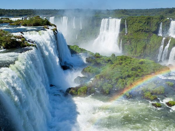 Majestic waterfalls cascade through lush green landscapes, with a vibrant rainbow arching over the misty river below.