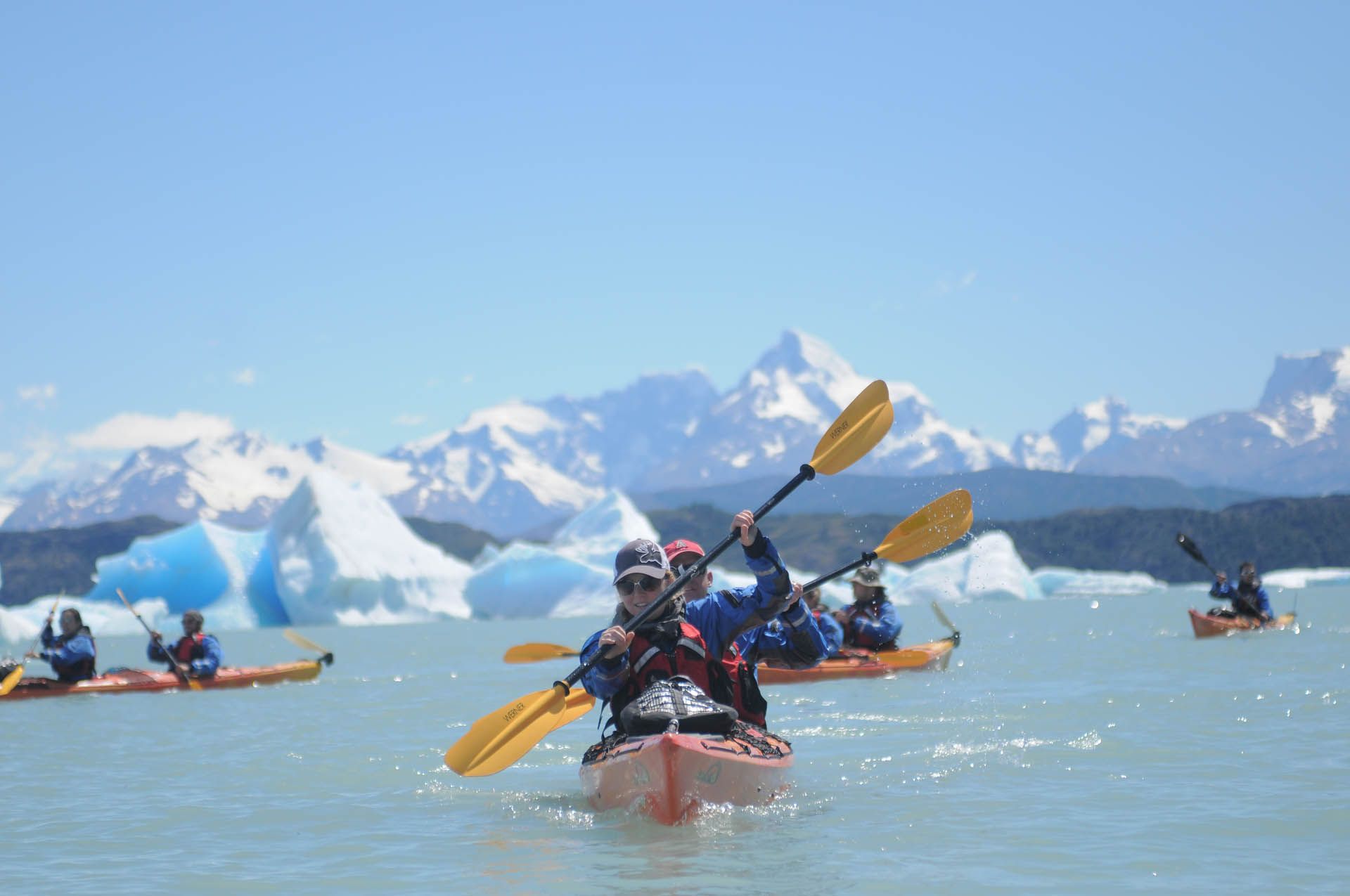 Kayakers paddle on calm water, past large blue icebergs and majestic snow-capped mountains under a clear sky at the front of Perito Moreno  Glacier