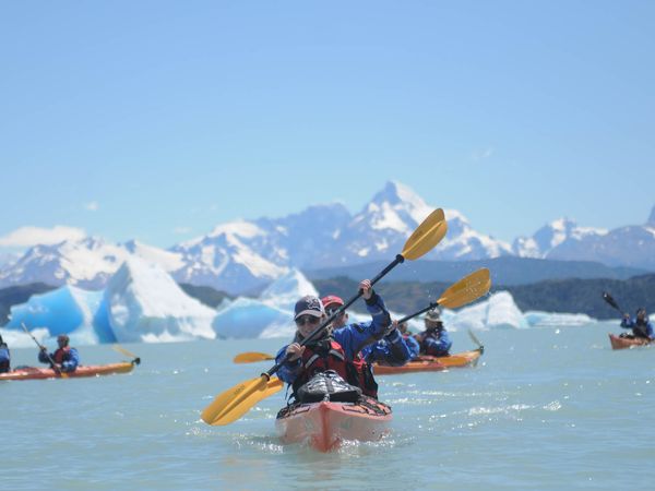 Kayakers paddle on calm water, past large blue icebergs and majestic snow-capped mountains under a clear sky at the front of Perito Moreno  Glacier