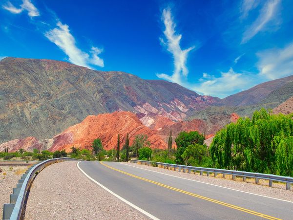 Scenic highway curves through colorful mountains in Argentina's Quebrada de Humahuaca with vibrant rock formations and native vegetation.