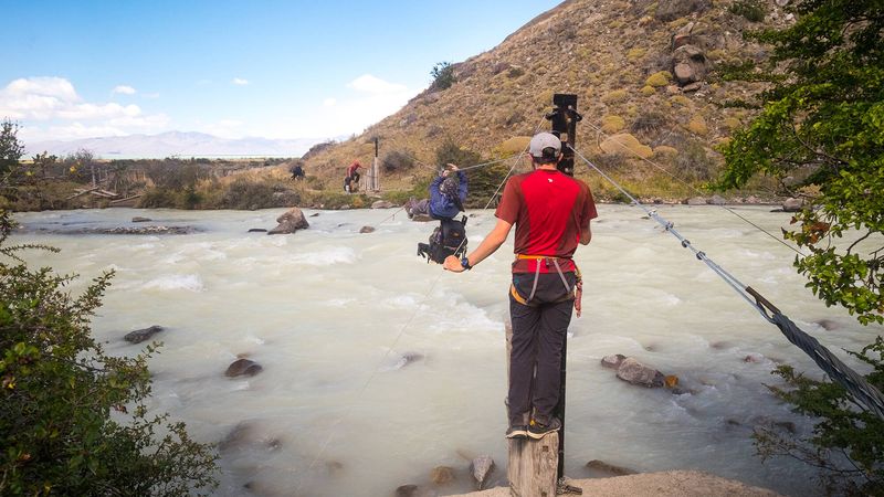 Adventure trekker crosses rope bridge over turquoise glacial river in El Chaltén, Patagonia.