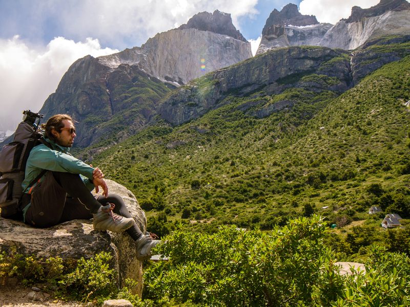 Hiker with backpack resting on rock overlooking lush valleys and dramatic granite peaks of Cuernos del Paine, Patagonia.