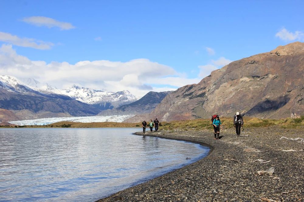 Hikers trek along a gravel beach beside turquoise glacial waters with Glaciar Viedma and snow-capped peaks in the background, El Chaltén.
