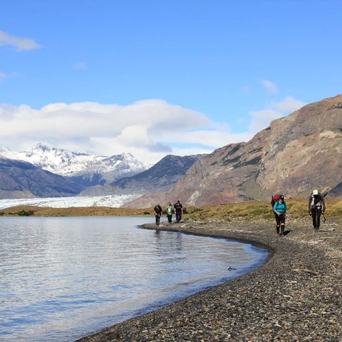 Hikers trek along a gravel beach beside turquoise glacial waters with Glaciar Viedma and snow-capped peaks in the background, El Chaltén.