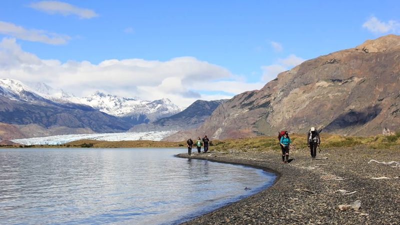 Hikers trek along a gravel beach beside turquoise glacial waters with Glaciar Viedma and snow-capped peaks in the background, El Chaltén.