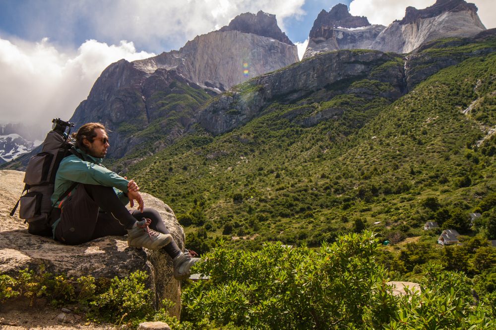 Hiker with backpack rests on rock overlooking lush valley with dramatic granite peaks of Torres del Paine, Chile.