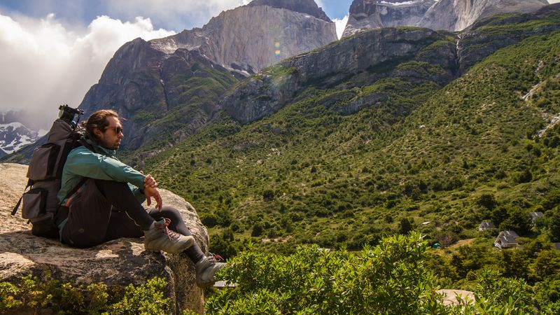 Hiker with backpack rests on rock overlooking lush valley with dramatic granite peaks of Torres del Paine, Chile.