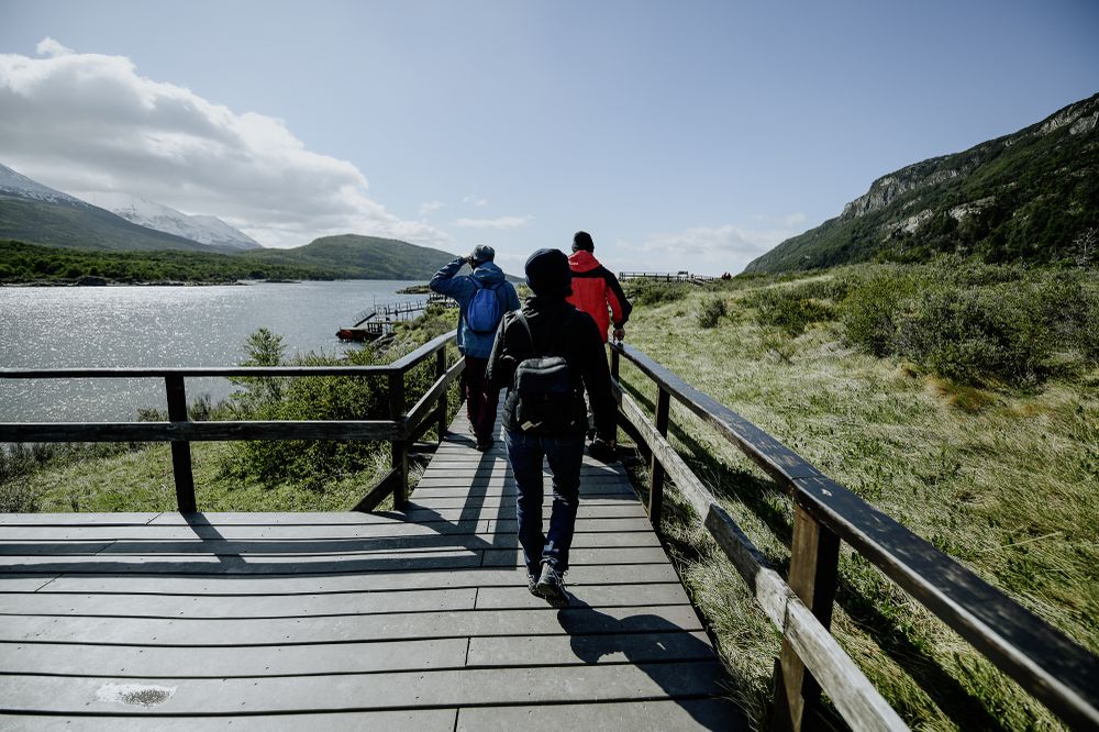 Hikers on a wooden boardwalk overlooking a pristine lake surrounded by mountains in Ushuaia National Park, Patagonia.
